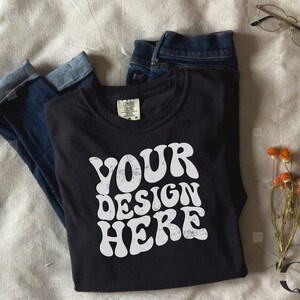 Peut inclure: Un t-shirt noir à manches longues avec le texte "YOUR DESIGN HERE" en blanc, en lettres de style rétro. Le t-shirt est plié et posé sur une surface beige clair avec un jean bleu et des fleurs séchées.