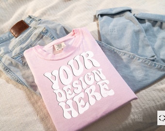May include: A pink t-shirt with the text "YOUR DESIGN HERE" in white, retro-style lettering. The shirt is folded and laid on a white surface with a pair of blue jeans.