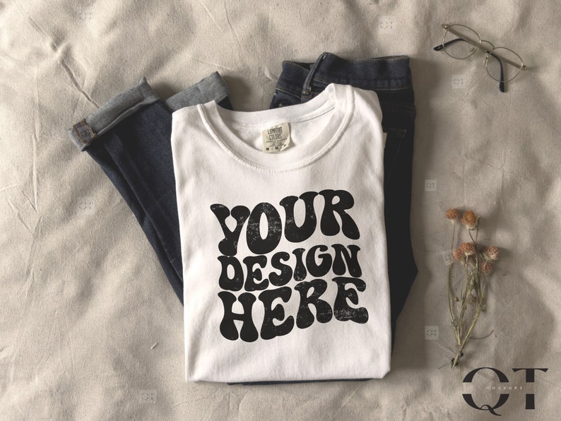 May include: A white t-shirt with the text "YOUR DESIGN HERE" in a retro font, folded on top of a pair of dark blue jeans. The image also includes a pair of eyeglasses and a small bouquet of dried flowers, all arranged on a neutral-toned fabric.