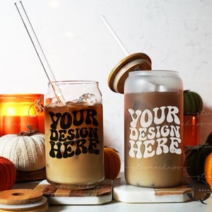 May include: Two glass tumblers with iced coffee, one clear and one frosted, each with the text "YOUR DESIGN HERE". A clear straw and a wooden lid are visible. The scene includes decorative pumpkins and a lit candle.