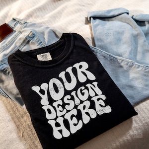 May include: A black t-shirt with a white graphic that says "YOUR DESIGN HERE" in a retro font. The shirt is folded and placed on top of a pair of blue jeans.
