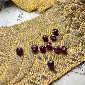 May include: A close-up of ten small, round, dark red glass beads on a gold fabric background. The beads are scattered on the fabric.