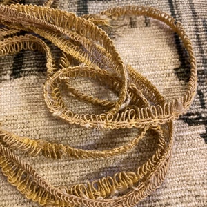 May include: Close-up of a length of gold-coloured decorative trim. The trim has a looped edge and is lying on a textured, neutral-toned surface. The trim appears to be made of a woven material.