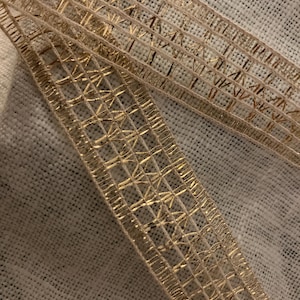 Puede incluir: Primer plano de una cinta metálica dorada con un patrón geométrico. La cinta es de color oro claro y tiene un diseño tejido. El fondo es una tela texturizada de color blanquecino.