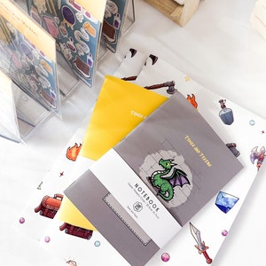 May include: A display of stationery items, including notebooks and sticker packs. The notebooks are gray and yellow, with a dragon illustration and the text "NOTEBOOK". Sticker packs feature adventure-themed icons. The items are arranged on a white surface.