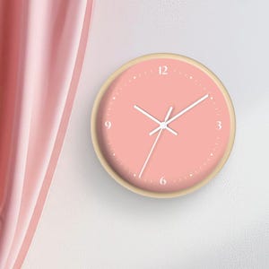 May include: A light pink clock with white numbers and hands. The clock has a wooden frame and is hanging on a white wall.