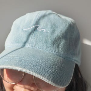 Könnte beinhalten: Hellblaue Jeans-Baseballkappe mit weiß gesticktem Wellenmuster. Die Kappe hat einen gebogenen Schirm und eine klassische Sechs-Panel-Konstruktion. Der Hut ist aus weichem, verblichenem Denim-Material.