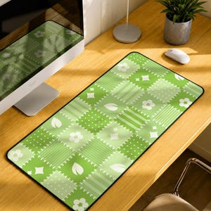 May include: A rectangular desk mat featuring a green patchwork pattern with alternating squares of white flowers, polka dots, stripes, and leaf shapes. The mat has a black stitched border and a smooth surface designed for computer mouse use.