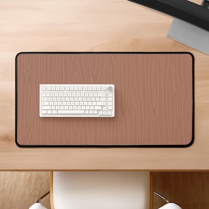 Op de afbeelding: Een bruine bureau mat met een houtnerf patroon. Een wit toetsenbord met een ronde knop ligt op de mat.