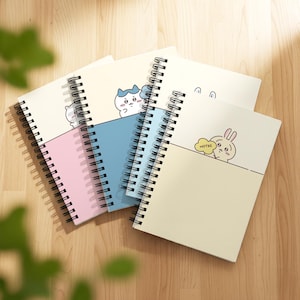 Könnte beinhalten: Eine Sammlung von Spiralnotizblöcken mit Cartoon-Tierillustrationen. Die Notizbücher haben Umschläge in Pastellfarben: Rosa, Blau und Creme. Jedes Notizbuch zeigt ein anderes Tierdesign, darunter eine Katze und ein Hase. Das Wort "NOTES" ist sichtbar.