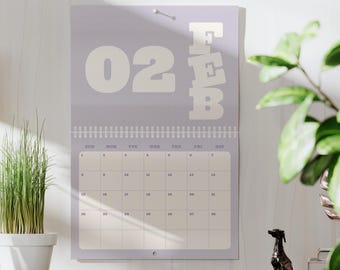 Può includere: Un calendario da parete di febbraio, con sfondo lilla chiaro che mostra il mese e la data in bianco. Il calendario è tenuto da una rilegatura a spirale argentata e una piccola puntina argentata. Una pianta in vaso e una statua di cane sono sullo sfondo.
