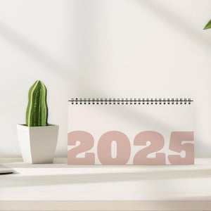 Puede incluir: Un calendario de escritorio rosa 2025 con fondo blanco y encuadernación de alambre negro. El calendario está sobre una superficie blanca.