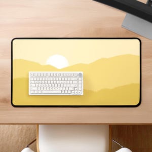 Op de afbeelding: Een gele bureau mat met een zonsondergang landschap ontwerp. Een wit toetsenbord met een ronde, grijze knop is op de mat.
