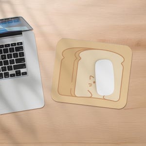 May include: A beige mouse pad with a cartoon illustration of a cat peeking out from behind a slice of bread. The mouse pad is on a wooden surface.