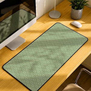 Puede incluir: Una alfombrilla de escritorio rectangular con un patrón repetitivo de pequeñas caras de conejo blancas delineadas sobre un fondo verde salvia. La alfombrilla tiene un borde negro cosido y está colocada sobre un escritorio de madera junto a un monitor.
