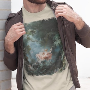 May include: A beige t-shirt featuring a vintage-style print of a painting depicting a woman on a swing in a lush forest setting. The painting is in shades of blue, green, and pink.