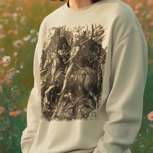 Puede incluir: Una sudadera beige claro con una ilustración en blanco y negro de una escena de batalla medieval. El gráfico representa caballeros a caballo y figuras en combate. La sudadera se lleva en un campo de flores.
