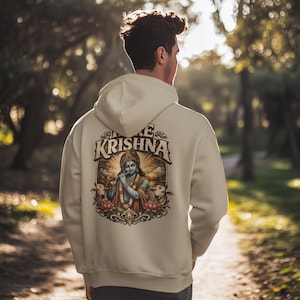Puede incluir: Sudadera con capucha color crema con un gráfico en la espalda. El gráfico presenta el texto "Hare Krishna" sobre una ilustración de una deidad azul tocando la flauta, rodeada de vacas y flores de loto. La sudadera tiene capucha.
