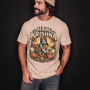 Puede incluir: Camiseta beige con las palabras "Hare Krishna" sobre una colorida ilustración de una deidad tocando la flauta, rodeada de vacas y flores de loto. La camiseta la lleva una persona con un gorro beige y vaqueros azules.