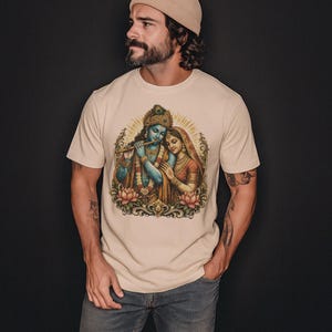May include: Beige t-shirt featuring a detailed illustration of two figures, one playing a flute, surrounded by lotus flowers and a golden halo. The figures are in shades of blue, red, and gold. The t-shirt is worn by a person.