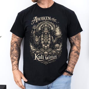 Puede incluir: Camiseta negra con un gráfico detallado de una deidad sosteniendo un tridente y un cuenco de fuego, rodeada de calaveras. El texto "Awaken the Kali Within" está sobre la imagen. El diseño es en tonos dorados y crema.