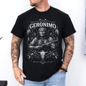 Op de afbeelding: Zwart t-shirt met een gedetailleerde illustratie van Geronimo, die een geweer vasthoudt, met de naam "GERONIMO" erboven. Het ontwerp bevat veren, een schedel en andere inheemse Amerikaanse afbeeldingen. Het t-shirt wordt door een persoon gedragen.