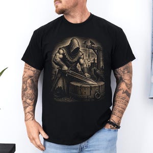 May include: A black short-sleeve t-shirt featuring a graphic print of a hooded figure in armor sharpening a large sword on a wooden stump. The scene includes a gallows structure with a noose, a raven perched nearby, and a dark castle silhouette.