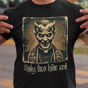 May include: Black t-shirt with a vintage-style image of a devil wearing a priest's collar. The text "Holy lies hide evil" is printed below the image.