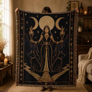May include: A rectangular woven throw blanket featuring a dark background with a central illustration of a figure wearing a crown and holding two flaming torches. Two dogs sit at the figure's feet, flanked by crescent moons and a full moon.