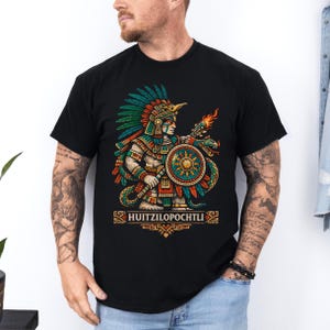 Peut inclure: T-shirt noir avec une illustration détaillée d'une figure aztèque tenant un bouclier et un serpent enflammé. Le design utilise des couleurs vives comme le vert, le rouge et l'or, avec le mot "HUITZILOPOCHTLI" en dessous.