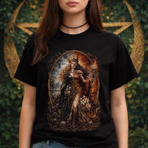 Puede incluir: Camiseta negra con una ilustración detallada de una mujer dividida en dos mitades, una oscura y otra ardiente. El diseño incluye calaveras, una corona y un báculo con una calavera. La obra de arte está enmarcada en un óvalo.