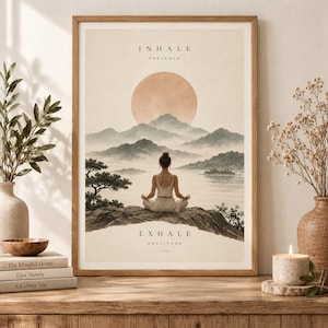 Op de afbeelding: Een ingelijste kunstprint met een persoon die mediteert op een rots met uitzicht op een meer en bergen onder een grote zon. De tekst INHALE PRESENCE staat bovenaan en EXHALE GRATITUDE onderaan. Het kleurenpalet bestaat uit gedempte beige, bruine en grijstinten.