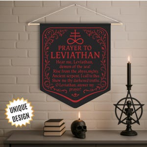 Pode incluir: Bandeira preta com texto vermelho que diz "ORAÇÃO A LEVIATÃ" e um poema. Uma vela de caveira preta, um castiçal de pentagrama preto com um esqueleto e uma pilha de livros estão em uma superfície cinza. Um adesivo "DESIGN ÚNICO" está à esquerda.