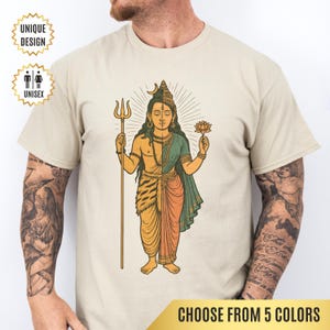 May include: Beige unisex t-shirt featuring a detailed illustration of a deity holding a trident and a lotus flower. The design is split vertically, with one side in orange and the other in green. The text "UNIQUE DESIGN" and "UNISEX" are also visible.