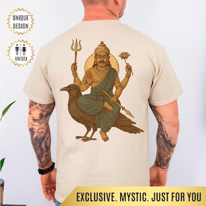 May include: Beige unisex t-shirt featuring a detailed illustration of a deity riding a crow, holding a trident, lotus flower, and a spear. The design includes a gold halo and is labeled "UNIQUE DESIGN" and "UNISEX". The bottom of the shirt reads "EXCLUSIVE. MYSTIC. JUST FOR YOU."