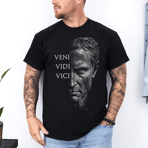 May include: Black t-shirt featuring a grayscale portrait of a man and the Latin phrase "Veni Vidi Vici" in white text. The design is on the front of the shirt. The shirt is made of a soft material.