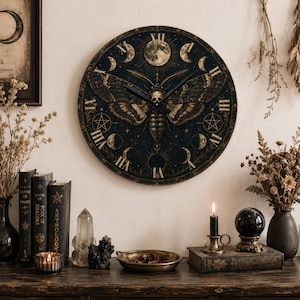 May include: A round wall clock featuring a dark blue and gold moth illustration at the center, surrounded by lunar phases and Roman numerals. The clock face displays a detailed moth with a skull pattern on its thorax, set against a starry background.