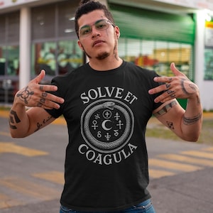 Puede incluir: Camiseta negra con el texto blanco "SOLVE ET COAGULA" que rodea un diseño circular con una serpiente, símbolos y una luna creciente. La camiseta está hecha de un material suave.
