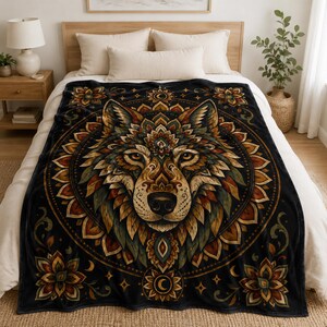 May include: A black throw blanket featuring a large, detailed illustration of a wolf head centred within a circular mandala pattern. The design incorporates warm tones of orange, gold, and muted green, with floral motifs and crescent moon symbols.