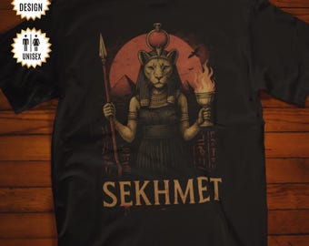 May include: A black t-shirt featuring a graphic of the Egyptian deity Sekhmet with a lion head, holding a spear and a flaming chalice. The design includes a red sun, pyramids, and hieroglyphics in the background, with the text SEKHMET below.