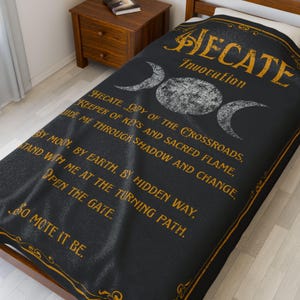 May include: Black blanket with gold text and a triple moon symbol. The text reads "Hecate Invocation" and includes a prayer to Hecate, the goddess of crossroads, keys, and magic. The blanket is on a bed.