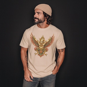Pode incluir: Camiseta bege com uma ilustração detalhada de um pássaro mítico dourado com as asas abertas. O pássaro tem detalhes em vermelho e verde. A pessoa usa um gorro bege e tatuagens.