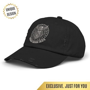 Puede incluir: Gorra de béisbol negra desgastada con un diseño circular de un hombre barbudo con casco alado y el texto "WISDOM THROUGH DEATH". La gorra tiene una visera curva y un aspecto envejecido.