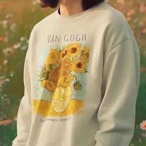 Puede incluir: Sudadera verde claro con una impresión de la pintura "Girasoles" de Van Gogh. La obra de arte representa un jarrón amarillo lleno de girasoles. El texto "VAN GOGH" está encima de la imagen, y "Sunflowers (1888)" debajo.