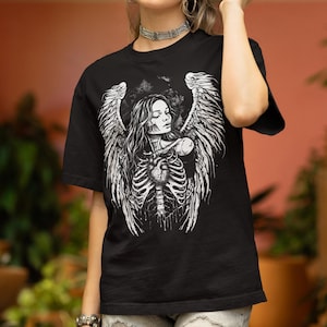 Puede incluir: Camiseta negra con un diseño gráfico blanco de una mujer con alas de ángel, una caja torácica y un corazón. El diseño tiene un tema gótico o de fantasía. La mujer tiene el pelo largo y los ojos cerrados. La camiseta es de material suave.