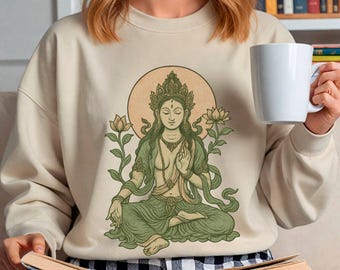 May include: Cream-colored sweatshirt featuring a serene illustration of a seated figure with a halo, lotus flowers, and intricate details in green and gold. The design evokes a sense of peace and spirituality. The sweatshirt is a comfortable, casual garment.