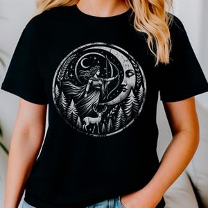 Może przedstawiać: Czarny T-shirt z białym nadrukiem graficznym przedstawiającym kobietę z łukiem i strzałami stojącą na rosnącym księżycu. Księżyc jest otoczony lasem i gwiazdami.