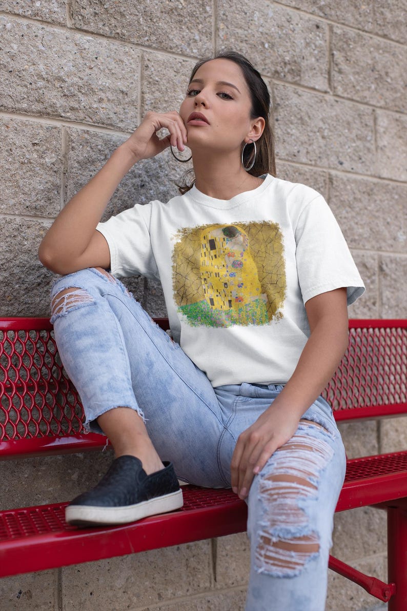 Distressed style t-shirt featuring Gustav Klimt painting The Kiss, rendered in faded gold and black tones on soft, white cotton fabric, suitable for art lovers.