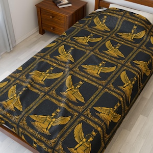 Könnte beinhalten: Eine schwarz-gold gemusterte Bettdecke mit einem sich wiederholenden Design einer ägyptischen Göttin mit ausgebreiteten Flügeln. Die Bettdecke ist über einem Einzelbett drapiert und zeigt die komplizierten Details des Kunstwerks.