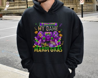 May include: Black hoodie with a graphic design featuring a purple video game controller, Mardi Gras beads, and the text "I paused my game for Mardi Gras." The design includes gold coins and a mask.
